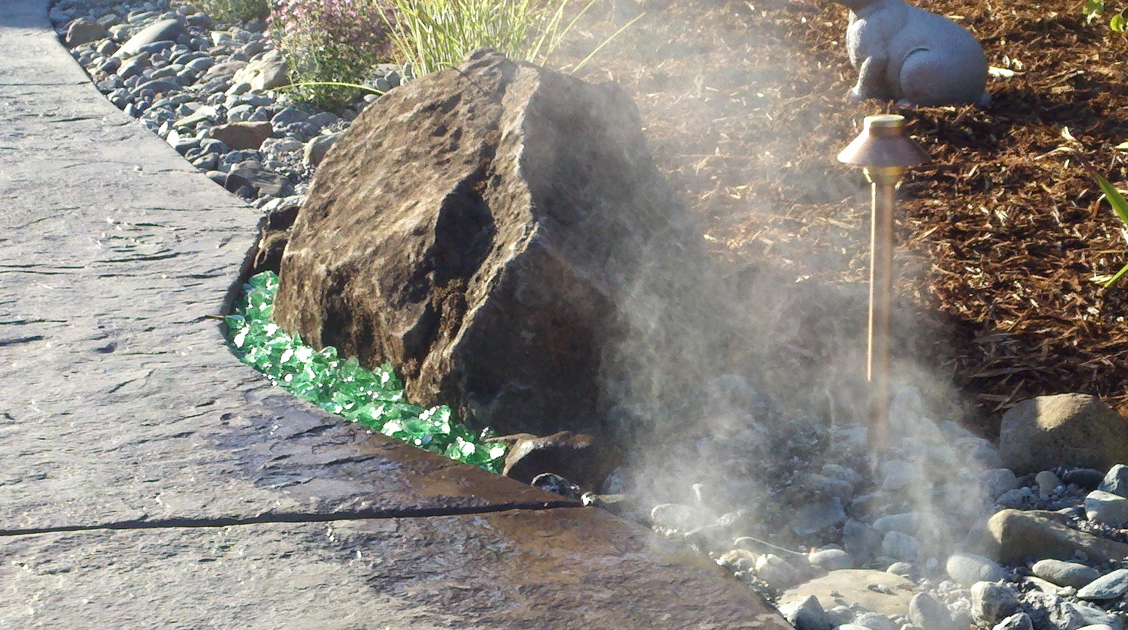 Landscape fogging nozzles releasing mist across a stone deck edge and boulder feature