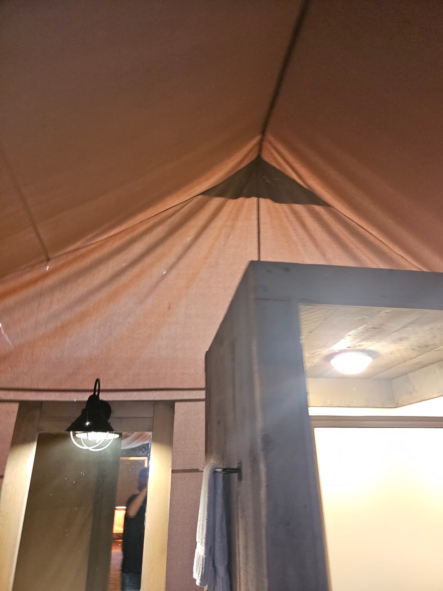 Inside a canvas glamping tent looking up at the floating main roof, ridge and wall panel above a framed doorway with a wall light