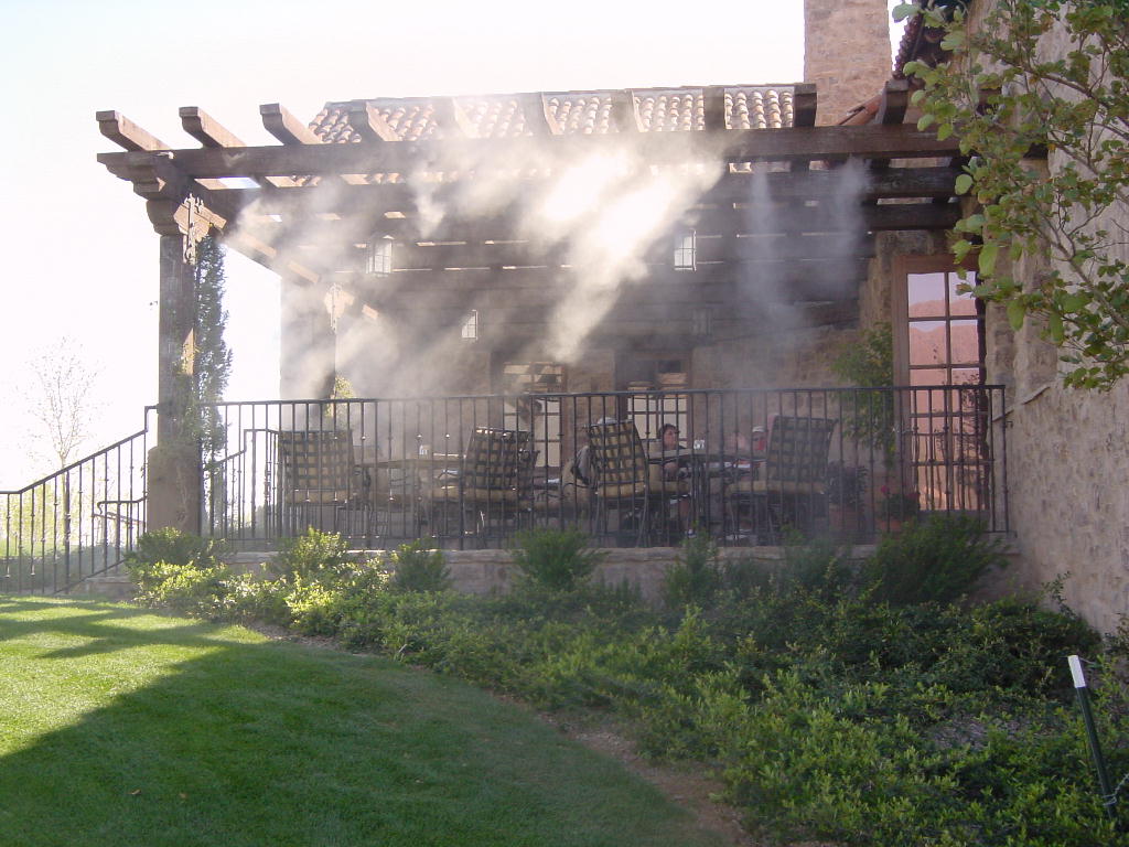 High-pressure misting nozzles fogging a shaded pergola patio where guests are seated at a table