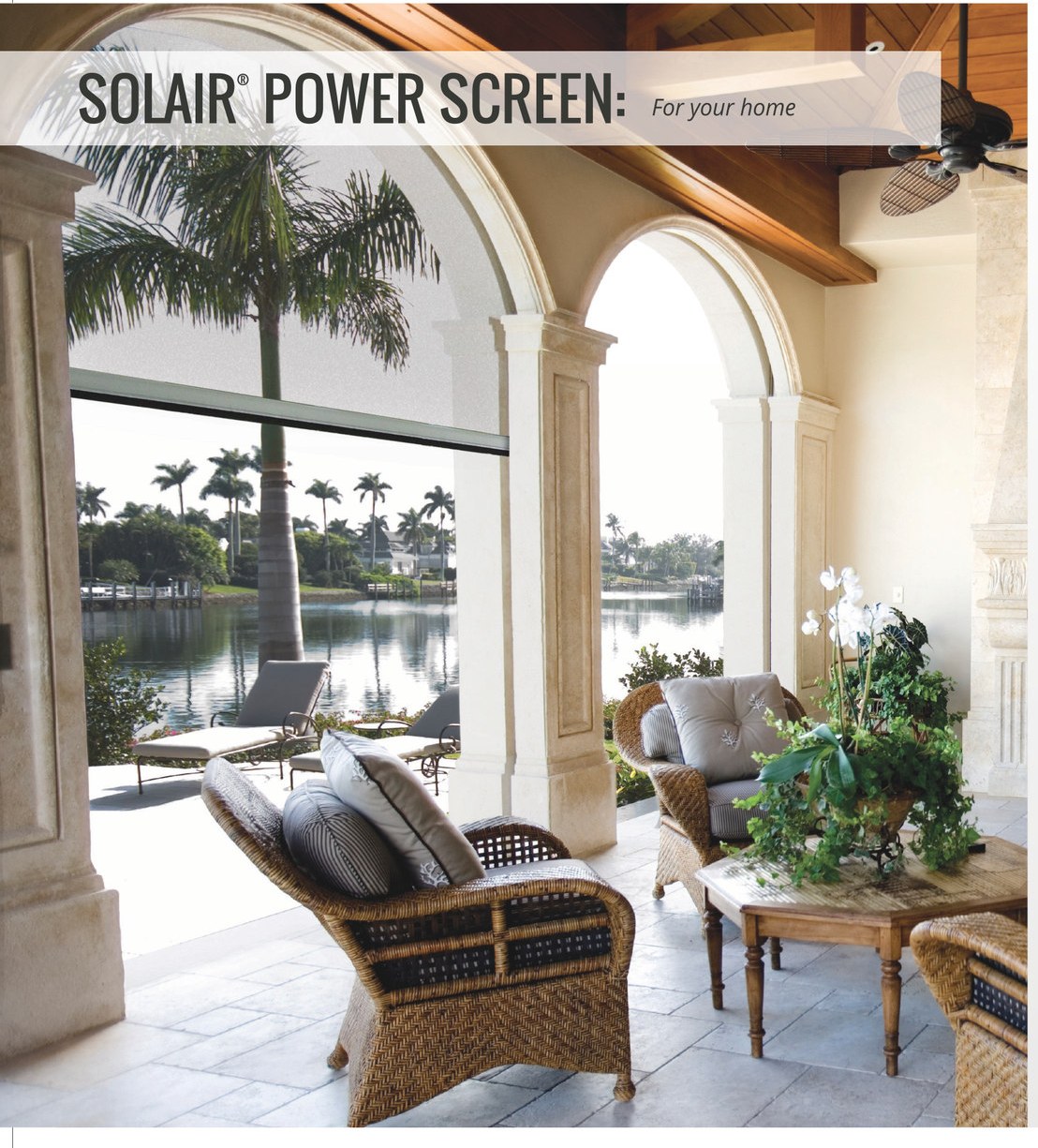 Solair® Power Screen lowered behind the arched openings of a waterfront lanai, shading wicker seating while the water view stays visible through the screen fabric