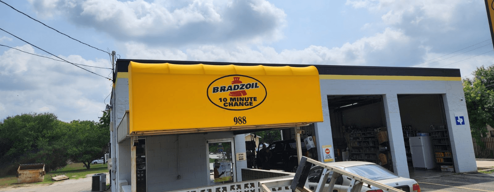 Bright yellow Sunbrella storefront awning with hand-painted Bradzoil 10 Minute Oil Change logo and 988 street number installed by Texas Made Shade