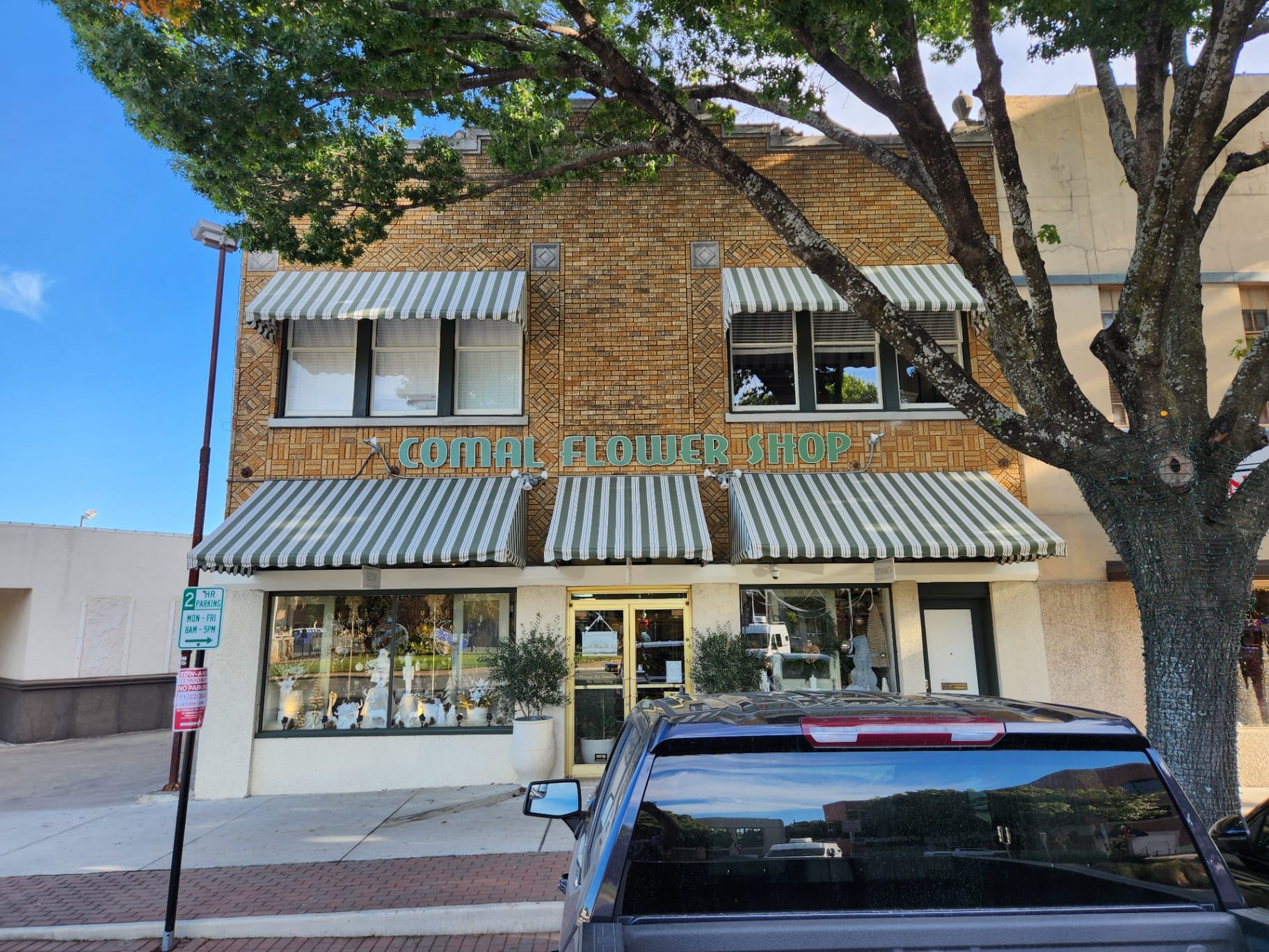 Front-elevation view of multiple custom green-and-cream striped Sunbrella commercial awnings installed by Texas Made Shade across the Comal Flower Shop brick storefront in New Braunfels, Texas