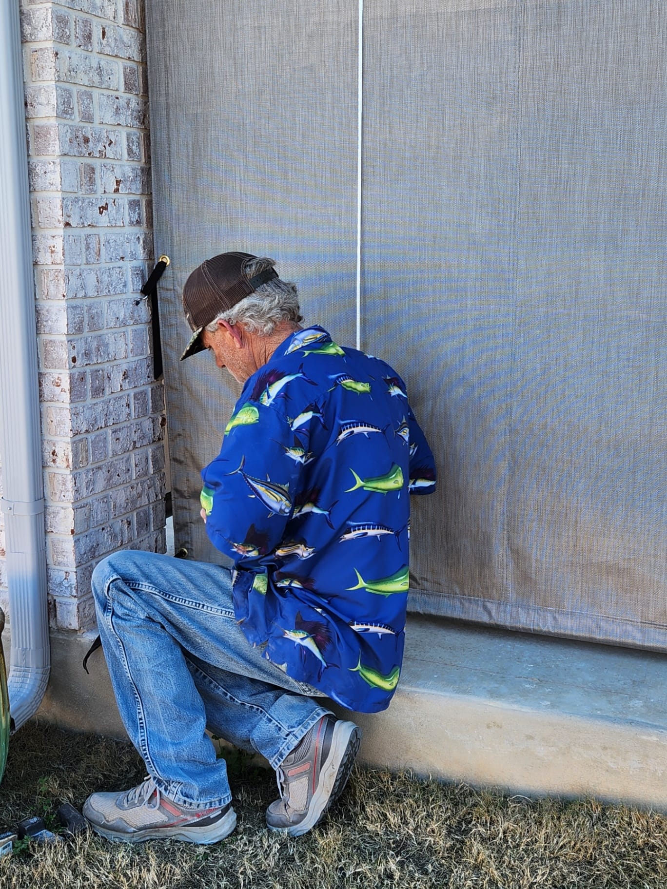 Texas Made Shade installer in a marlin-print shirt and ball cap kneeling on a Central Texas back patio while fastening a tan Phifertex mesh drop curtain to the brick column with a brass turn-button and pull cord