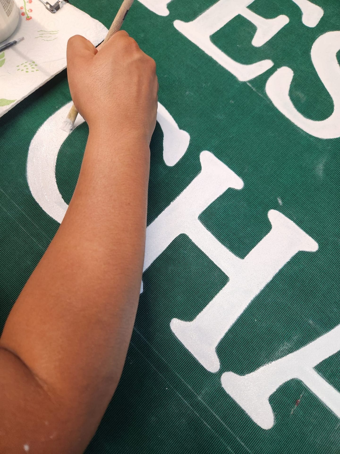Texas Made Shade artist hand-painting a white block-letter business logo onto deep green Sunbrella commercial awning canvas in the Seguin shop