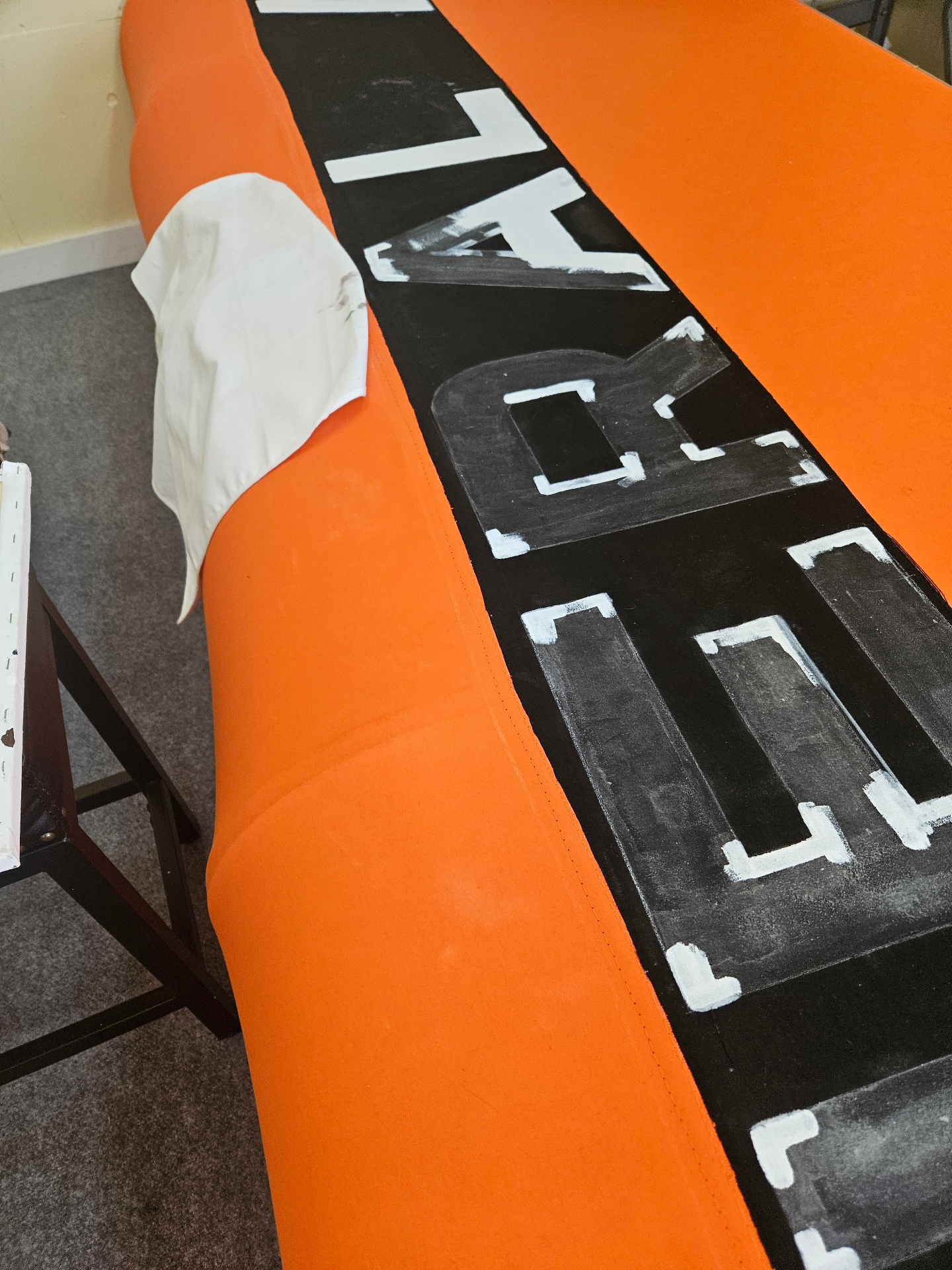 In-progress hand-painted black block lettering with white outline being applied by Texas Made Shade onto a bright orange Sunbrella commercial canopy laid out on sawhorses in the Seguin shop