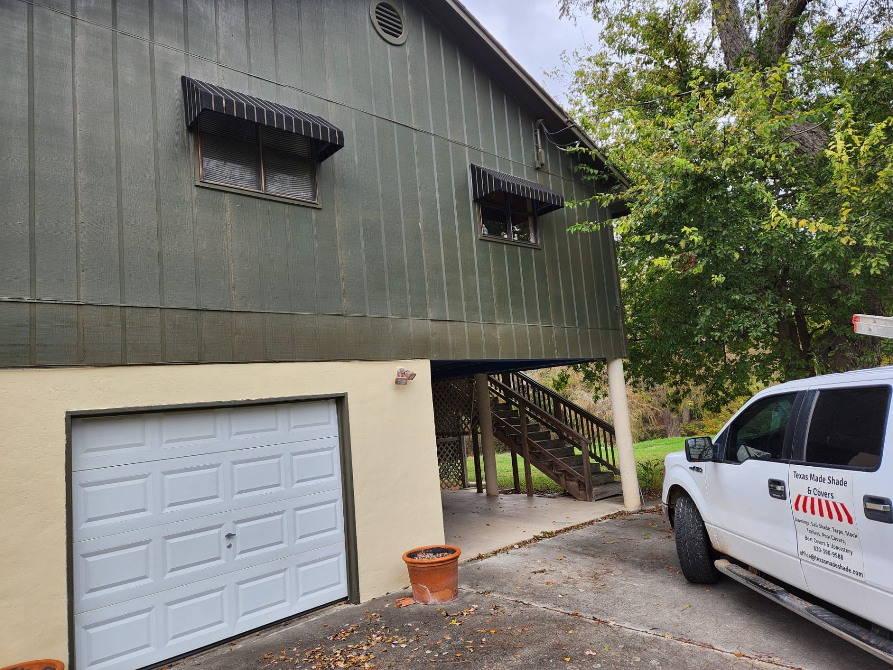 Pair of custom black-and-cream pinstripe Sunbrella window awnings fabricated and installed by Texas Made Shade over the upstairs windows of a green board-and-batten Texas home, with the Texas Made Shade & Covers work truck parked in the driveway