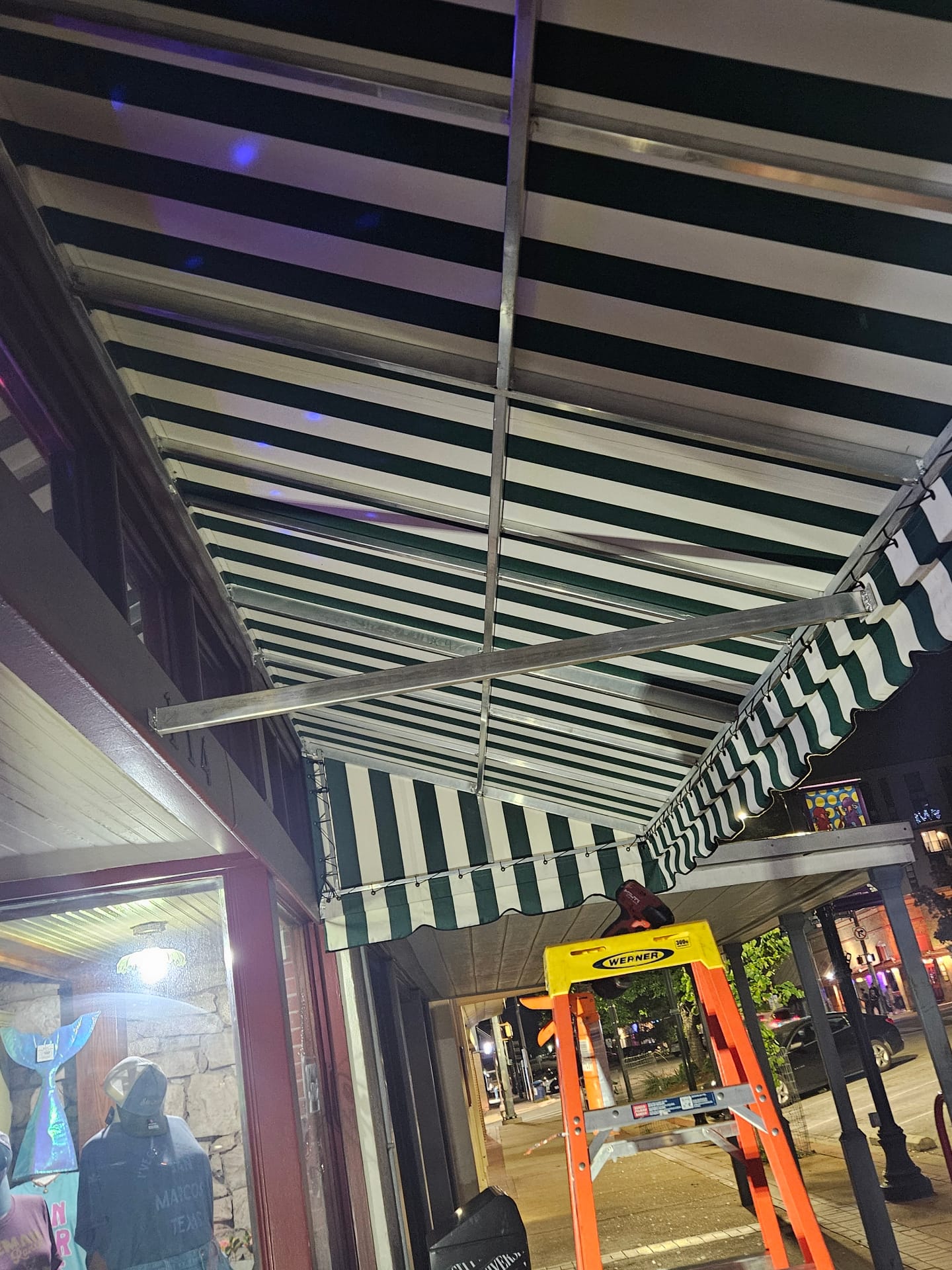 Underside view of a Texas Made Shade welded aluminum awning frame with green and white striped Sunbrella canvas stretched tight over a downtown San Marcos storefront