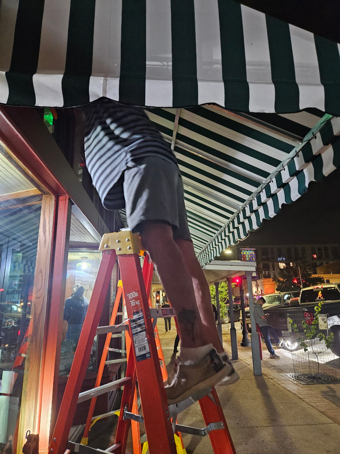 Texas Made Shade installer on a ladder fitting a custom green and white striped canvas awning to a welded aluminum frame at The San Market storefront in downtown San Marcos at night