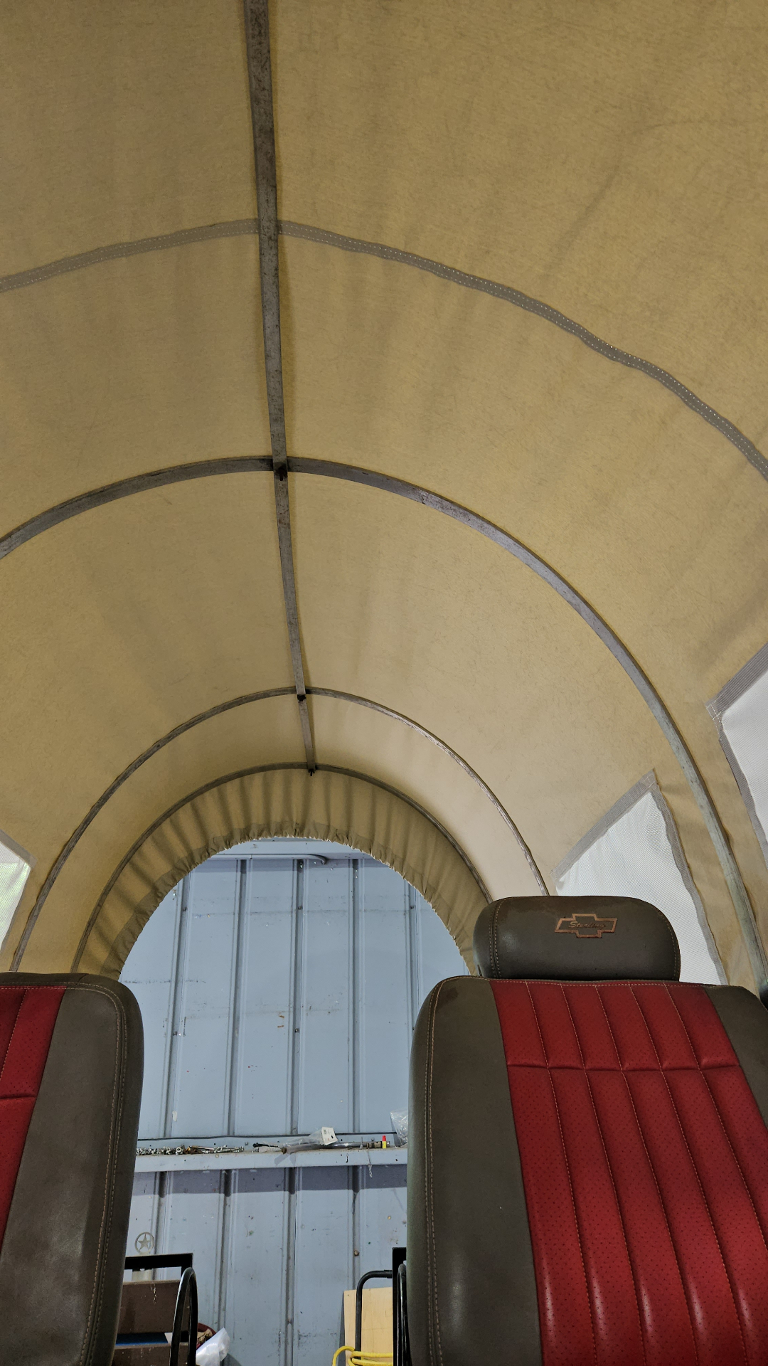 Interior view from inside a custom Texas Made Shade covered wagon — heavy canvas top stretched across steel bow frame with reinforced binding tape, framing two red-and-brown leather Chevrolet bench seats and an arched rear opening