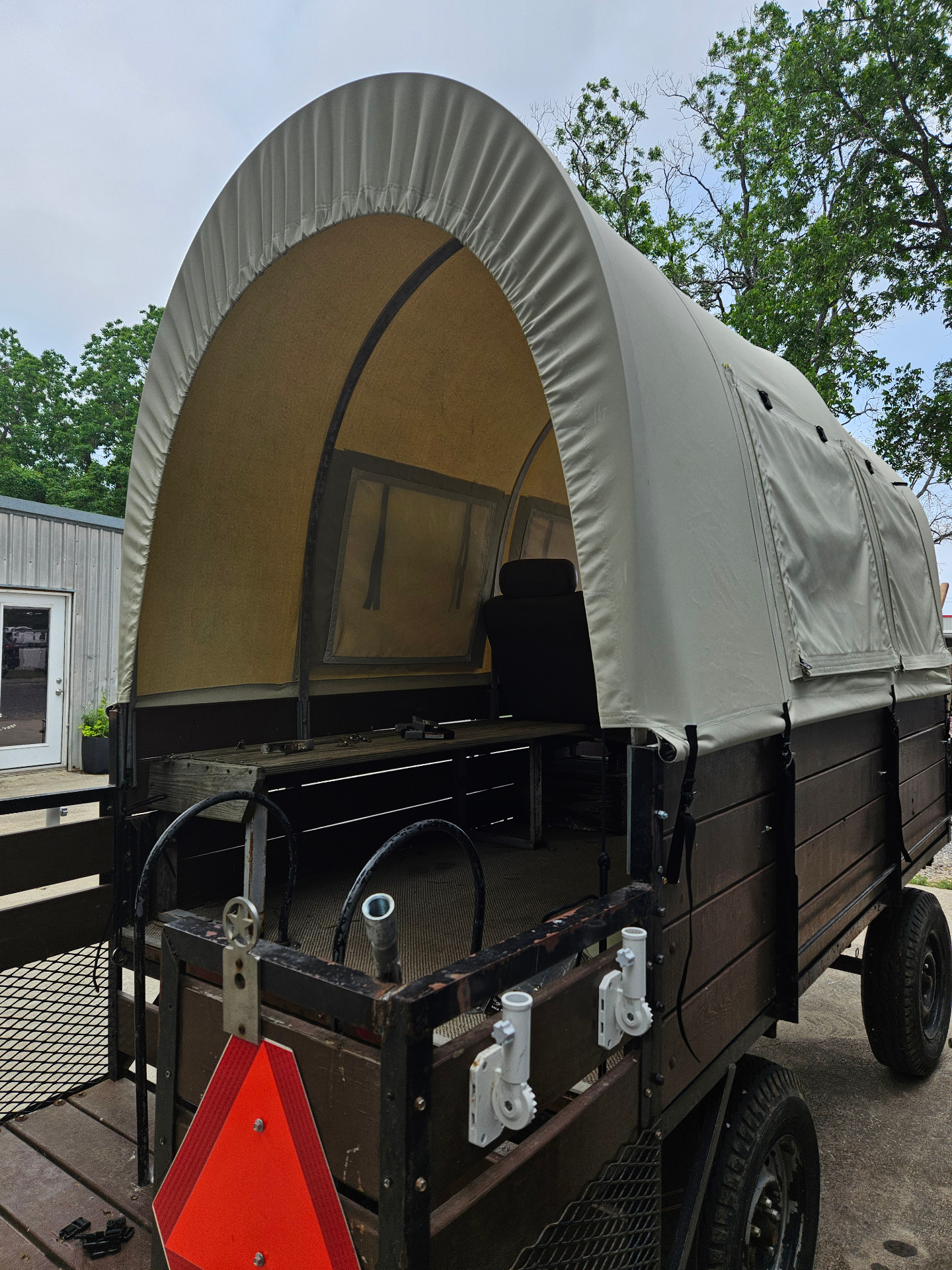 Custom natural-canvas covered wagon top fabricated by Texas Made Shade over a wood-sided utility trailer chassis, showing the rear arched opening, gathered drawstring closure, and bow-framed canvas roof
