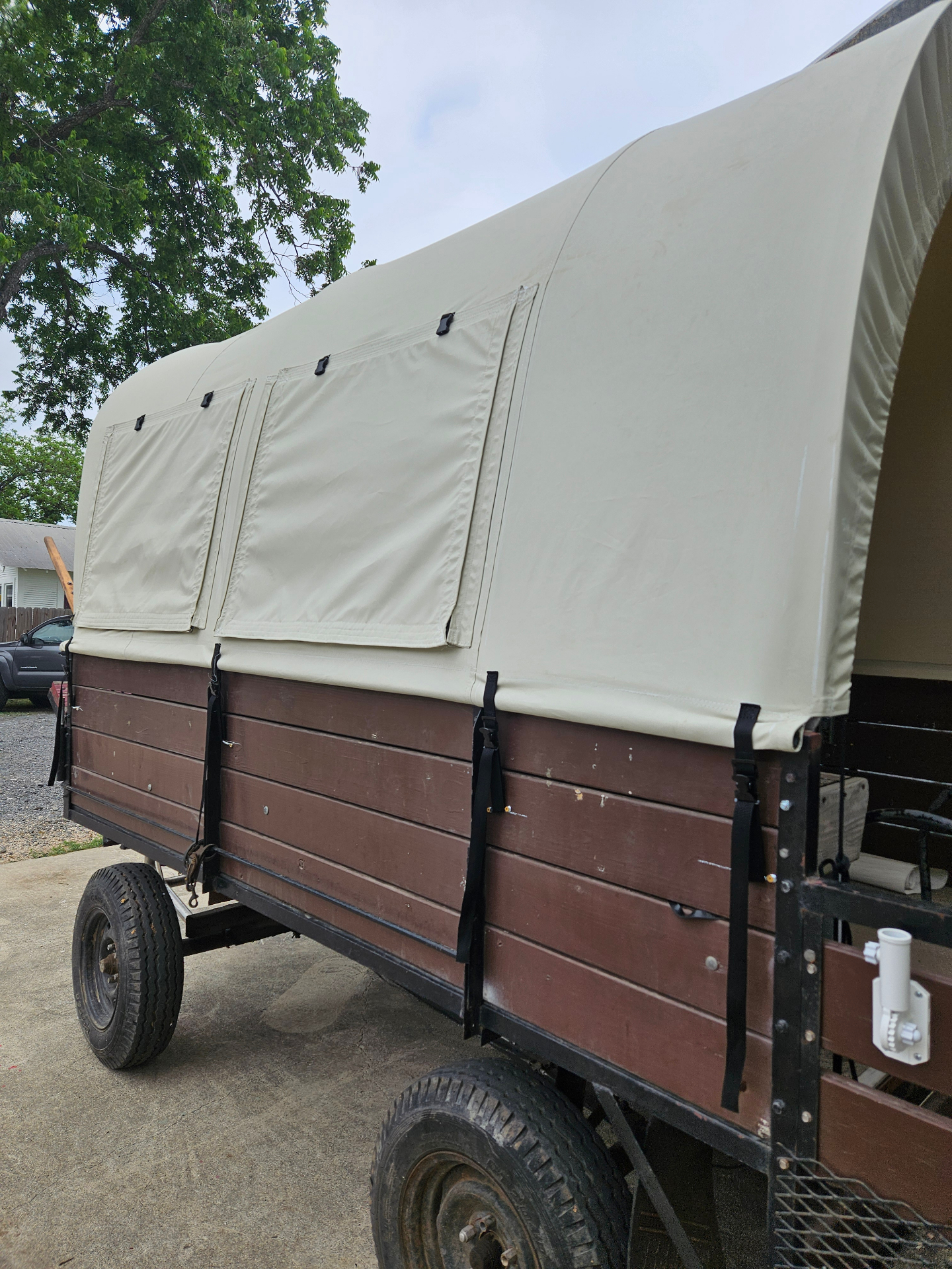 Side view of a custom Texas Made Shade covered wagon top on a brown wood-sided trailer, showing two roll-up screen window flaps with buckle closures and black webbing tie-down straps running down each bow to the trailer frame