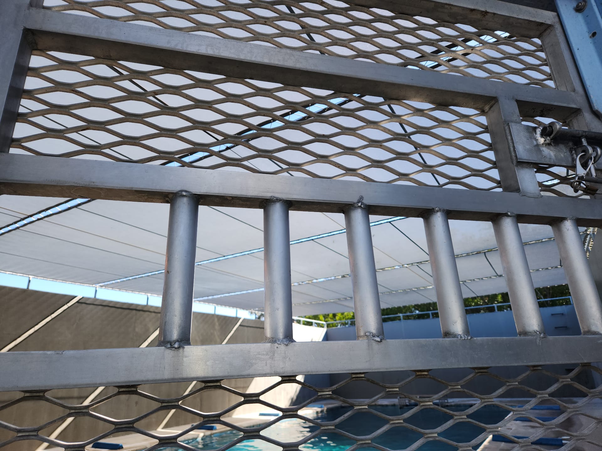 Close-up of a welded aluminum gate with expanded metal mesh panels and welded tube bars fabricated by Texas Made Shade, framing a tensioned white shade sail over a commercial pool deck