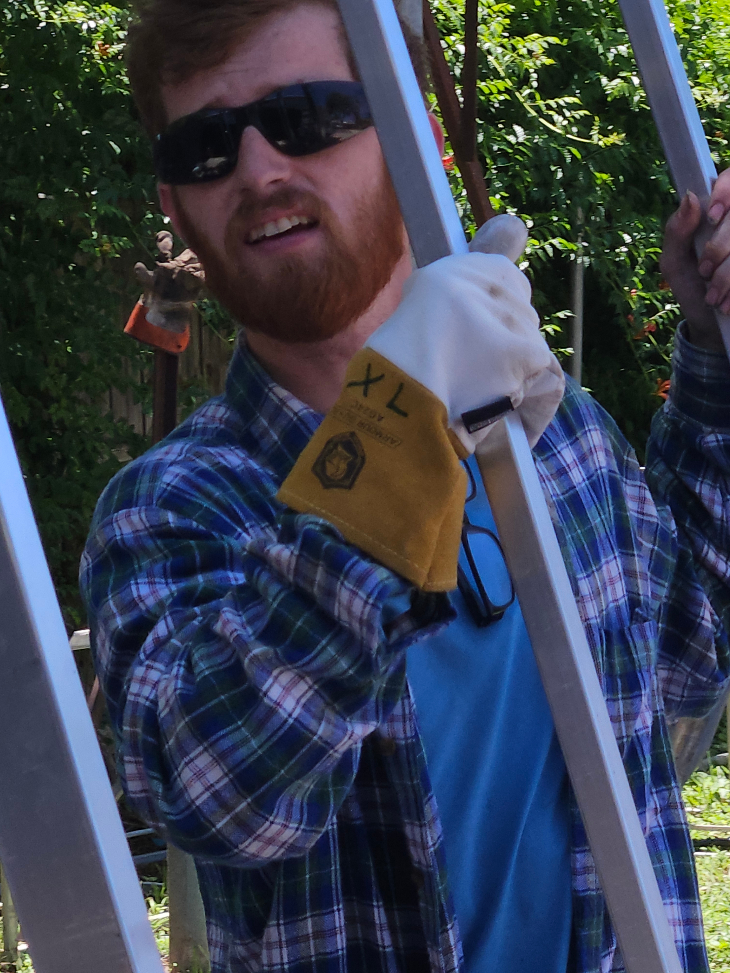 Close-up of a Texas Made Shade fabricator in welding gloves and safety glasses standing inside a freshly built aluminum awning frame at the Seguin, TX shop, gripping the vertical tubing during a final fit-check